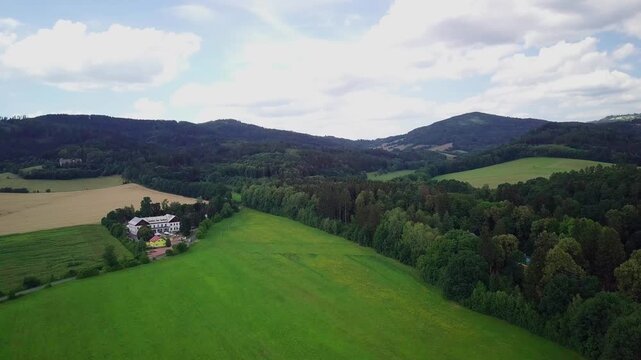 Drone flies over a green meadow and forest edge panning to reveal a large white building on a hill below forested ridges near the spa town of Velk&eacute; Losiny, Czechia.