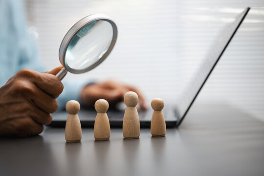 A magnifying glass hovers over wooden figures in a people review concept, with the magnifying glass directing attention along the small lineup as candidate search is symbolized beside an open laptop.