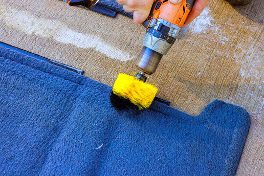 Hand holds drill with brush to clean blue rug cleaning floor mat on floor.