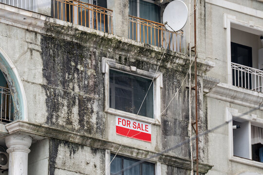 Heavily water-damaged residential building facade with red and white text sign "For Sale" in Batumi, hard to sell private property in Georgia due to poor house quality and poverty, dampness and mildew