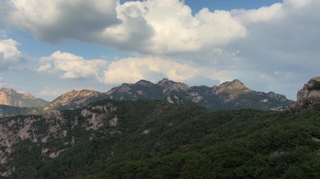 Forested mountain ridge with exposed rock faces under large white clouds