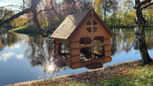 Wooden bird feeder shaped like a cabin by a calm river, rustic birdhouse with seeds in autumn park