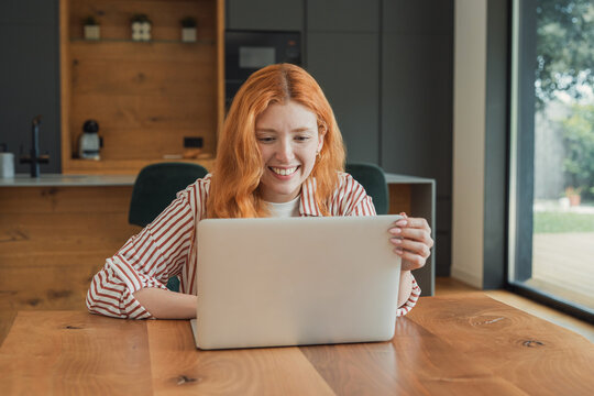 Young woman with red hair happily engaging with her laptop, creating a productive home office environment in her modern kitchen while managing remote work