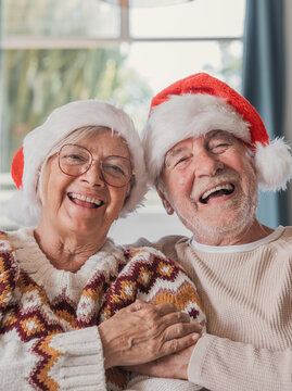 Elderly couple laugh together in Santa hats and patterned winter sweaters, holding hands at home. Their joyful Christmas celebration captures warm, heartwarming love and happy holiday connection.