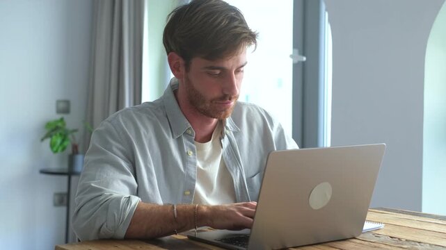 Man types on a laptop at a wooden home desk before pausing to look aside. Static footage shows focused freelance work, computer use, productivity, and independent employment indoors.
