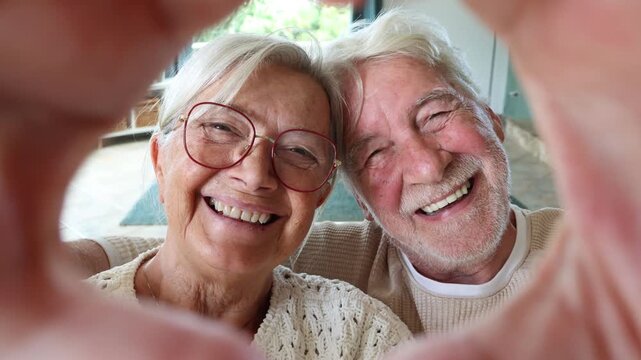 Joyful senior couple smiling and looking at the camera while making a heart shape with their hands. Happy caucasian retired people showing love, affection and sharing beautiful moments at home