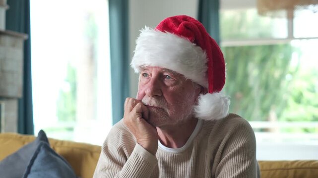 Elderly man misses family on Christmas morning as he sits alone on a sofa, resting his chin on his hand. His downcast gaze conveys holiday loneliness, separation and concern for senior wellbeing.