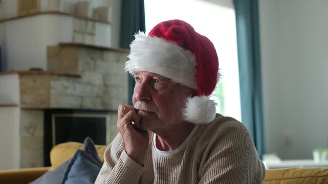 Portrait of a pensive and lonely senior man wearing a santa hat, feeling sad and abandoned while sitting alone on a couch at home during the christmas holiday season