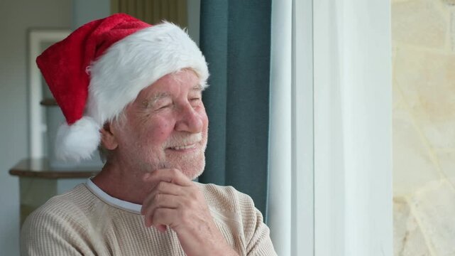 Senior man in a Santa hat gazes out the window, lifts his hand to his chin and smiles softly beside sheer curtains. Natural light and his beige sweater frame a reflective Christmas scene of solitude.