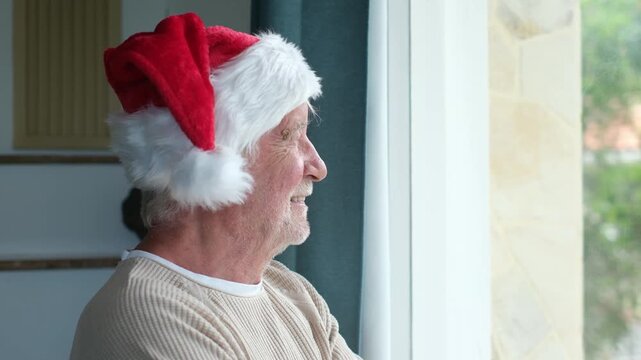 Cheerful senior man wearing a red santa hat smiles while looking out the window, anticipating the arrival of family for christmas holidays and feeling the joyful spirit of the season