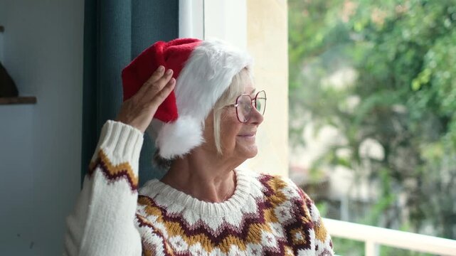 Cheerful senior woman in a festive sweater and a red santa hat smiling while looking out the window, enjoying the christmas holiday season with a contemplative and happy expression