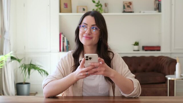 Woman smiles at the camera, looks up thoughtfully, then types on her smartphone while sitting at a table in her bright home. Her changing gaze shows mobile browsing, digital connection and technology.