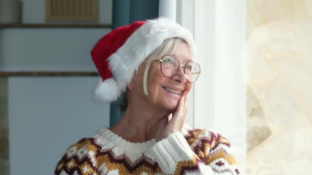 Happy senior woman in a santa hat and festive sweater looking thoughtfully out a window, smiling and waving, enjoying the christmas holiday season with warmth and nostalgia at home