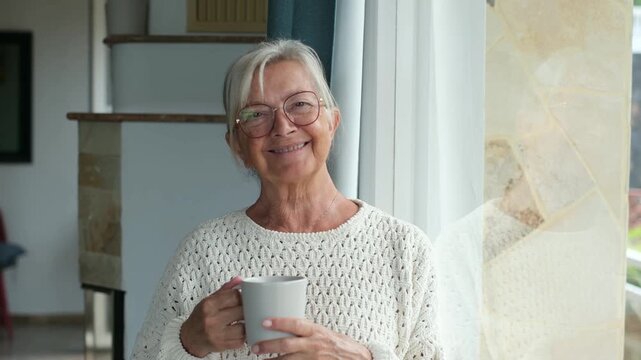 Beautiful older woman standing by the window holding a coffee mug, thoughtfully looking outside and then smiling at the camera, enjoying a moment of quiet relaxation at home