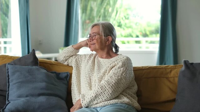 Thoughtful elderly woman with grey hair and glasses sitting on a cozy couch at home, looking away and smiling gently while enjoying a quiet moment of peaceful relaxation and contentment