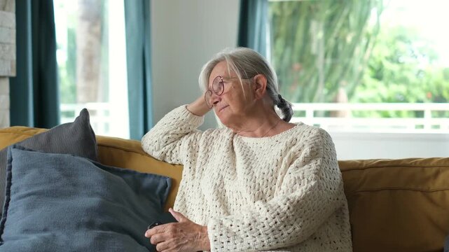 Elderly woman watches television from a yellow sofa, shifting from a pensive pose to a relaxed lean in her bright living room. She holds a remote control as soft natural window light fills the room.