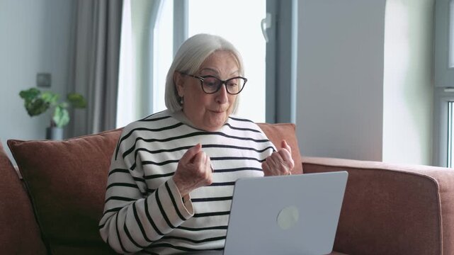 Overjoyed senior woman in glasses looking at her laptop screen, feeling happy about receiving good news, celebrating a victory or achievement while sitting on the sofa in her living room