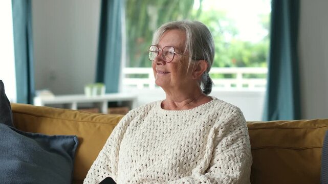 Peaceful elderly woman with grey hair and glasses smiling while resting on a comfortable couch in her living room, enjoying a movie or tv show with a look of contentment and happiness