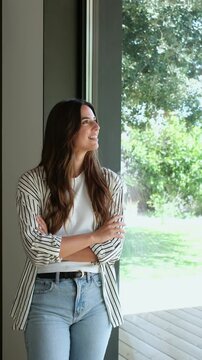 Confident young businesswoman with arms crossed smiling while looking out the window of a modern office, enjoying the view of the green garden and feeling optimistic about her future career