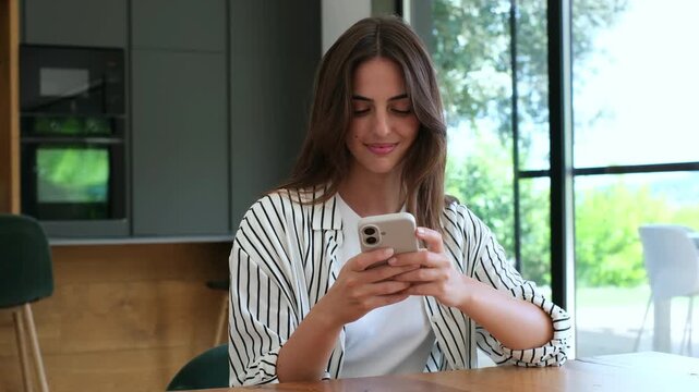 Smiling young woman types messages on a smartphone while seated at her kitchen table. Her cheerful phone use in a sunlit home captures casual texting, digital connection and daily communication.