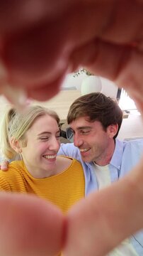 Adorable young couple making a heart shape with their hands while taking a selfie video at home. This point of view shot captures their joyful smiles and loving embrace in a cozy apartment setting