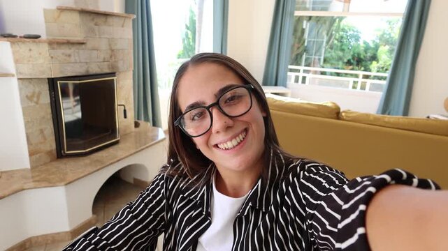 Woman records a selfie vlog in a sunlit living room, speaking to the camera before waving hello. Handheld POV captures a warm greeting, direct communication and casual content creation at home.