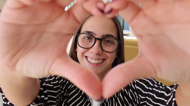 Charming young woman with glasses smiling at the camera while making a heart shape with her hands, expressing love and affection from a first-person perspective in a modern living room