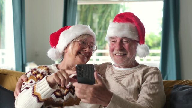 Senior couple share a smartphone and laugh together in Santa hats on a sofa. Elderly woman taps the screen as her husband holds her close, enjoying Christmas technology and holiday connection.