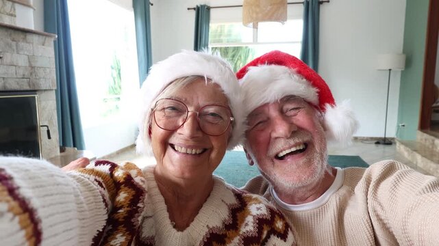 Cheerful senior couple wearing santa hats smiling and laughing while taking a selfie at home, enjoying the christmas holidays together and spreading festive joy in a video call