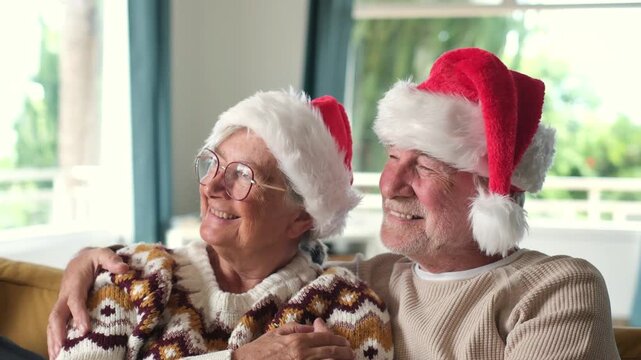 Elderly couple cuddle on a sofa in Santa hats, smiling as the man holds his partner close. They look offscreen in a bright living room, sharing affectionate Christmas time and holiday romance.