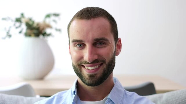Portrait of a handsome young caucasian man with a beard and blue eyes sitting on a couch at home, looking at the camera and smiling with confidence in a modern living room interior