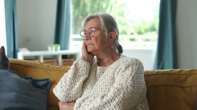 Thoughtful mature lady sitting alone on the couch, feeling pensive and melancholy while contemplating her life, loneliness, and health problems. Elderly woman experiencing solitude at home