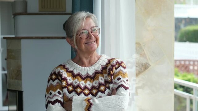 Elderly woman stands with folded arms by a large window and smiles toward the daylight. Her quiet pose communicates retirement, independent living, comfort, healthy aging and wellbeing at home.