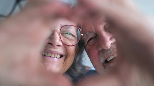Elderly couple lower their hand heart to reveal smiling faces indoors. Close-up static shot follows the man and woman looking at the camera, sharing a joyful gesture of love and companionship.