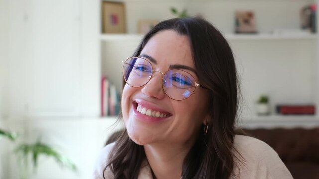 Woman in round glasses smiles, laughs, and rests her chin on her hand while looking aside at home. Her attentive expression and warm indoor setting convey relaxed listening, joy, and everyday.