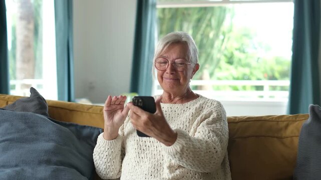 Senior woman waves hello, taps her smartphone and laughs during a video call on a couch in her bright living room. Her cheerful conversation shows digital connection and mobile communication at home.