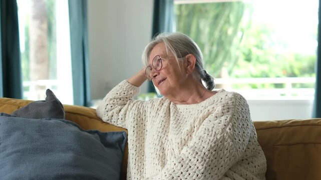 Thoughtful and serene elderly woman with grey hair and glasses smiling and relaxing comfortably on a sofa at home, enjoying her quiet retirement and feeling content in her living room