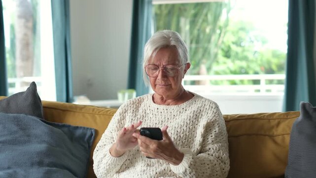 Elderly caucasian woman sitting on a sofa at home, getting stressed and anxious while reading bad news on her mobile phone, feeling worried and covering her mouth with her hand in shock