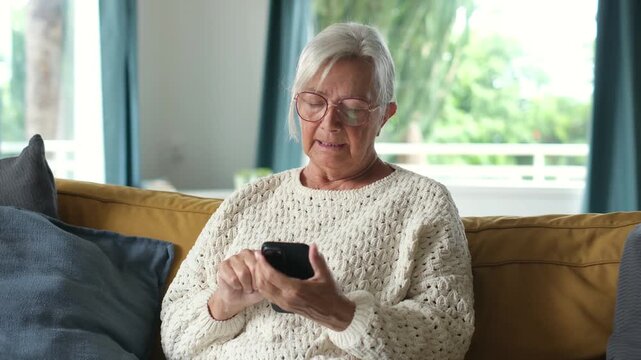 Senior woman taps and scrolls on a smartphone while seated on a yellow living room sofa. Grey-haired woman uses mobile technology at home, focused on browsing, digital literacy and online.