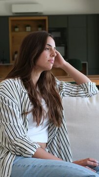 Woman sits alone on a sofa, rubs her temple and closes her eyes through headache discomfort. Her troubled expression conveys anxious isolation, mental fatigue and a need for stress relief.