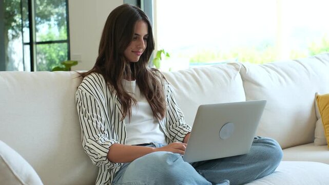 Beautiful smiling woman using a laptop computer, sitting on a comfortable couch in a modern living room. A freelancer working from home, she is typing on her keyboard and browsing the internet