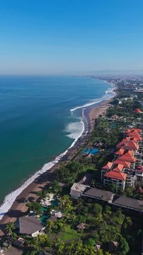Vertical high-angle drone shot of luxury beach resort with tiled roof villas, swimming pools, palm trees, and ocean waves. Great for social media reels and travel stories. Semenyak beach.