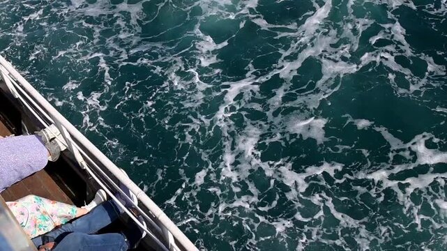 Top view from ferry in Istanbul, Turkey, looking down at dark blue Bosphorus water with white foam and passenger feet visible near the deck edge. Dynamic maritime travel scene with cpy space.