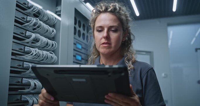 Female Server Room Specialist Studies Changing Server Metrics on Tablet, Seriously Analyzing Performance Data and Considering Possible Causes of Unusual System Behavior and Network Instability.