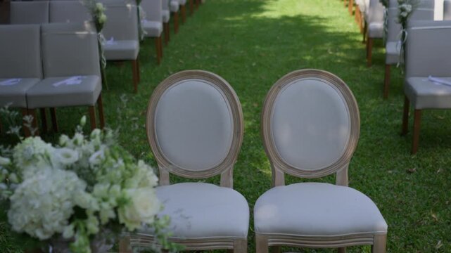 Garden ceremony setup with two upholstered medallion seats for the couple facing rows of guest seating on grass, white hydrangeas and roses beside the aisle.