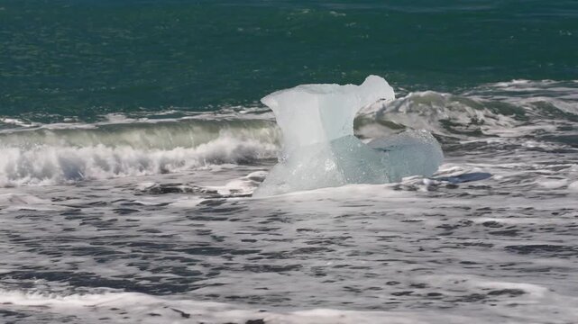 Ocean waves wash over chunks of blue glacial ice scattered across Iceland's black sand beach, creating a dramatic contrast of ice, volcanic sand, and Atlantic surf.