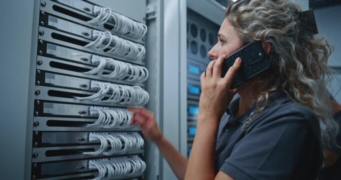 Female IT Specialist Consults Colleague by Smartphone While Inspecting Cables Connected to Server Rack, Coordinating Technical Support and Troubleshooting to Restore Reliable Network Infrastructure.