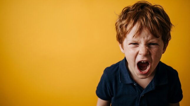 Angry young boy with red hair and freckles screaming with wide open mouth against yellow background