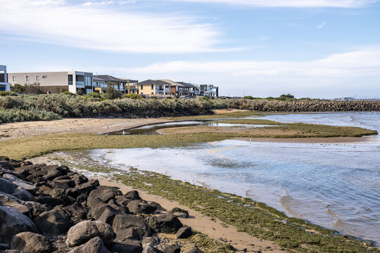 Low tide near Wyndham Harbour in Werribee South, Melbourne, Australia, featuring basalt rock sea walls, tidal mudflats with seaweed, coastal vegetation, and modern waterfront residential houses.