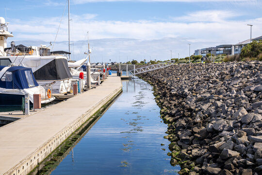 Moored boats in marina at Wyndham Harbour, with a calm water channel, a rock breakwater, a gangway to the pontoons and residential houses in the background. Werribee South, Melbourne, Australia